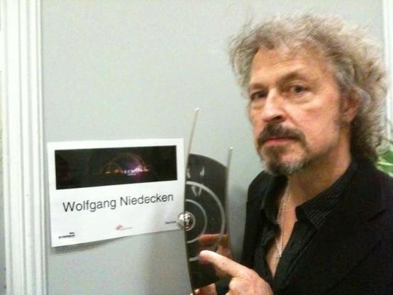 Das Bild zeigt einen Mann, der vor einer Tür steht, an der ein Schild mit der Aufschrift „Wolfgang Niedecken“ angebracht ist. Auf dem Schild ist ein Logo mit einem abstrakten Design und einem dunklen Hintergrund zu sehen. Die Person hält einen Preis oder eine Trophäe in der Hand, die aus einem glänzenden Material besteht und ein modernes, geschwungenes Design mit kreisförmigen Elementen hat. Die Tür und Wand im Hintergrund sind hellgrau, und die Szene wirkt wie in einem Backstage- oder Veranstaltungsbereich aufgenommen.