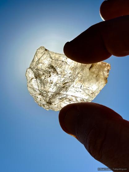 A close-up photograph of a translucent mineral or rock held delicately between two fingers. The rock's crystalline structure is illuminated by sunlight, revealing intricate fractures and inclusions within. Against the vibrant blue sky, the mineral glows, showcasing its transparency and internal patterns.