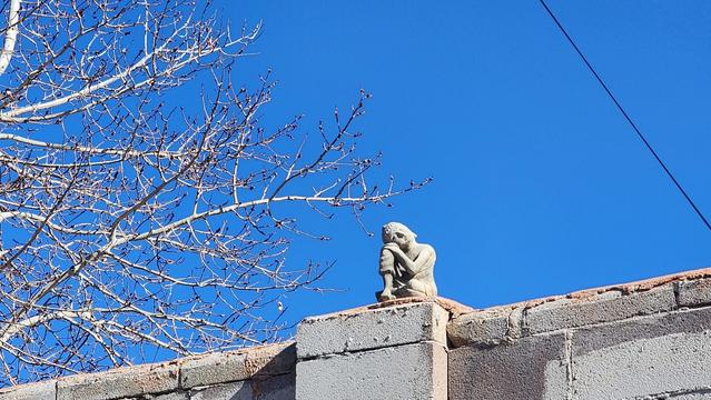 color photo of concrete humanoid Buddha figure sitting on a block wall. A clear blue sky and early spring tree in the background.
