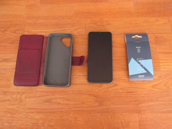 Fairphone 4 on a wooden table. To the left there is a red leather flip case. To the right a replacement battery still in its card board wrap.