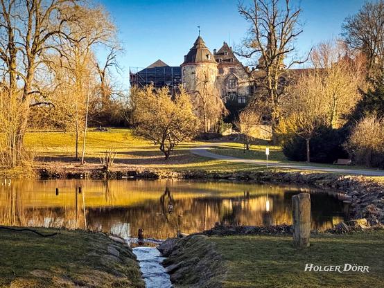 Blick auf das Schloss Laubach im warmen Licht des Frühlingsanfangs. Im Vordergrund ein kleiner Bach, der in einen ruhigen Teich mündet. Die Wasseroberfläche spiegelt die Bäume und das Schloss. Sanftes Sonnenlicht taucht die Landschaft in goldene Töne, erste Frühlingszeichen sind in der Parklandschaft sichtbar.