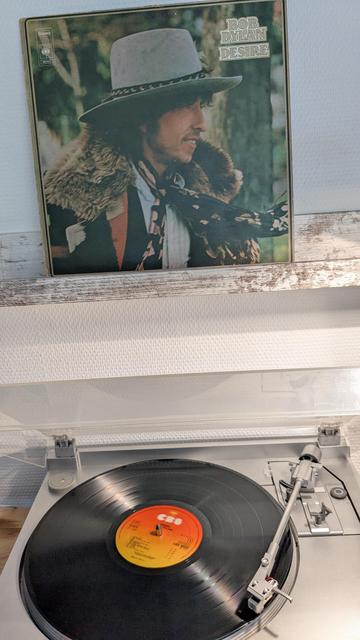 Das Bild zeigt einen Plattenspieler, auf dem eine Vinyl-Schallplatte abgespielt wird. Die Schallplatte hat ein orangefarbenes Label mit dem CBS-Logo. Im Hintergrund ist das Albumcover von Bob Dylans „Desire“ zu sehen. Das Cover zeigt eine Person mit einem breiten Hut, einem Schal und einem Pelzmantel in einer natürlichen Umgebung. Der Plattenspieler hat eine silberne Oberfläche und eine transparente Abdeckung. Die Szene wirkt gemütlich und nostalgisch, passend zur analogen Musikwiedergabe.