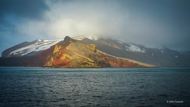 The chiselled landscape of Antarctic in glowing sunlight.  The hills have hues of copper, brown, yellow and green.  Patches of snow can be seen under drifting clouds that transition from white to blue / black toward the edges of the frame.