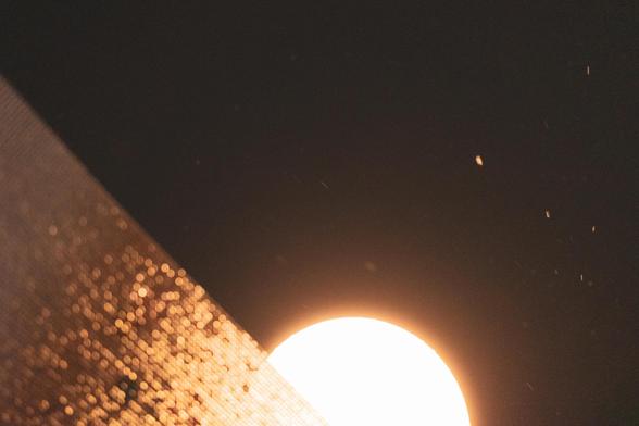 The image shows a bright circular light source, possibly the moon or a lamp, against a dark background. There appears to be a textured surface or material in the foreground, lit from below, creating a diagonal line across the image, with small particles or specks floating in the air.