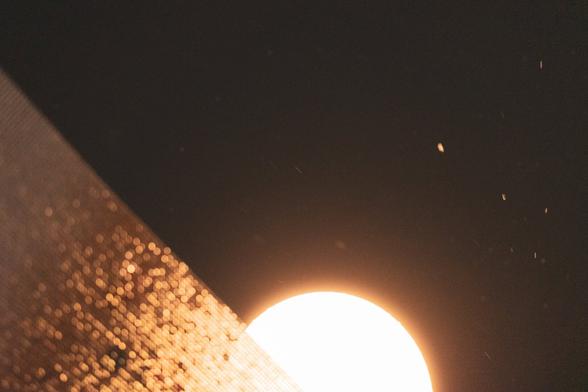 The image shows a bright circular light source, possibly the moon or a lamp, against a dark background. There appears to be a textured surface or material in the foreground, lit from below, creating a diagonal line across the image, with small particles or specks floating in the air.