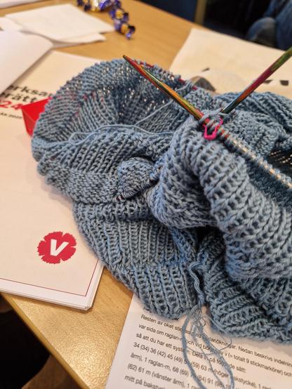 En bunt med papper med partilogga (röd nejlika med vitt v) En blågrå stickning på rundsticka. Några godispapper. Allt på en bordsskiva i ljust trä.