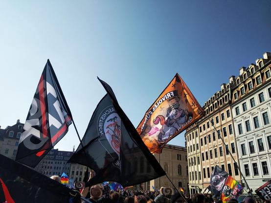 Fahnen der antifaschistischen Demo auf dem Neumarkt Dresden vor blauem Himmel, im Hintergrund ist der sich in Gang setzende Nazi-Aufmarsch zu sehen