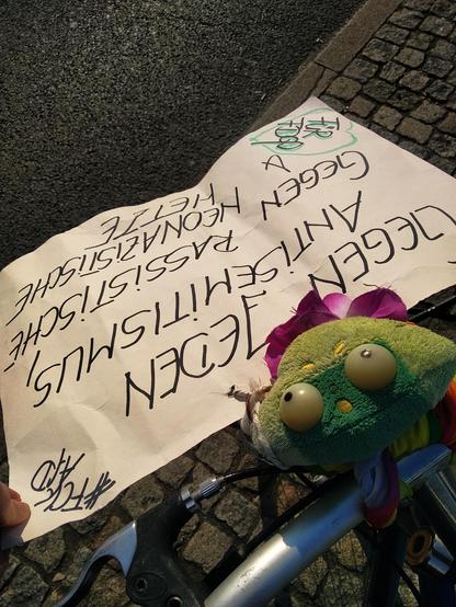 Blick auf den Lenker meines Fahrrades an dem Quaki mit Regenbogentuch und Blume hängt, davor das Plakat mit der Aufschrift "Gegen jeden Antisemitismus, gegen Rassismus, gegen Neonazistische Hetze. Für FDGO. #fckAfD"