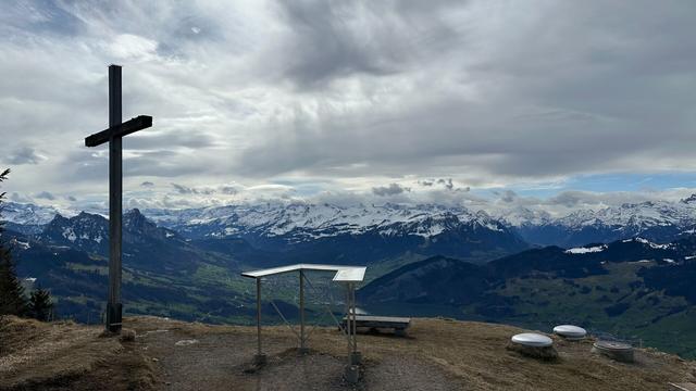 View from the Wildspitz looking south towards the Swiss Alps. A cross and a guide to the mountains are in the foreground on a hill covered in brown grass. The valley below is green and the sky is full of clouds.