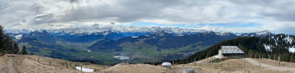 Panorama looking south towards the Swiss Alps from the Wildspitz. The Berggasthaus is in the foreground on the right. The sky is almost completely clouded over with a narrow strip of blue on the horizon.