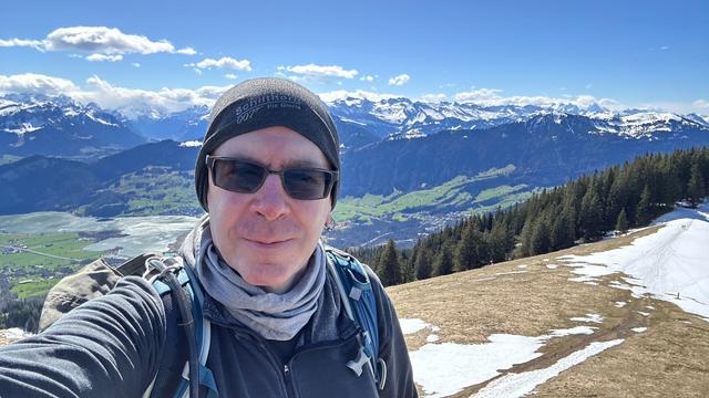 Selfie of me, a middle-aged white guy in sunglasses taken against a backdrop of a green valley and snow-covered mountains in the distance.