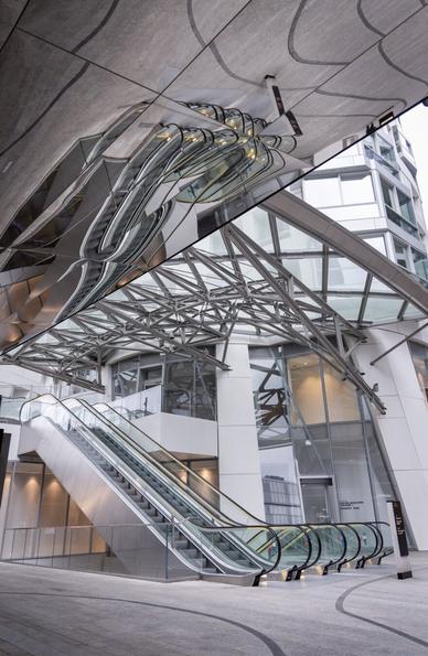 A modern architectural space featuring an escalator leading to an upper level, surrounded by glass and metal structures. The ceiling has a large mirrored surface that reflects the escalators and structural beams, creating a distorted and artistic visual effect. The building's exterior consists of glass windows and white paneling, with a geometric metal framework supporting a glass canopy. The scene has a futuristic and urban aesthetic, with clean lines and a sleek design.