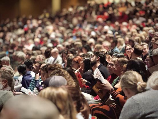 A sea of people sitting in auditorium chairs faces frame left toward a theatre stage under tungsten house lights.