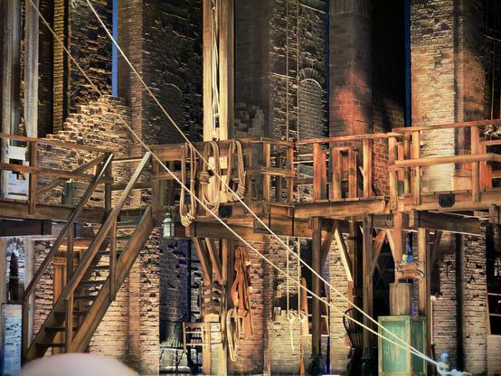 A theatre set representing a 1700s American industrial alleyway stands on stage, comprised of wooden steps leading to wooden rafters, brick walls, and numerous wooden boxes/bundles of rope.