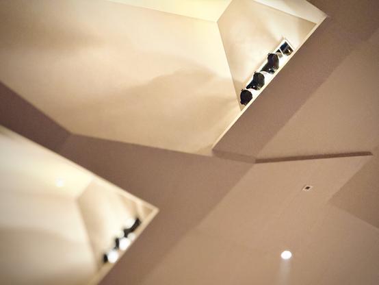 Black stage lights sit recessed in a large angular alcove of a white auditorium ceiling.