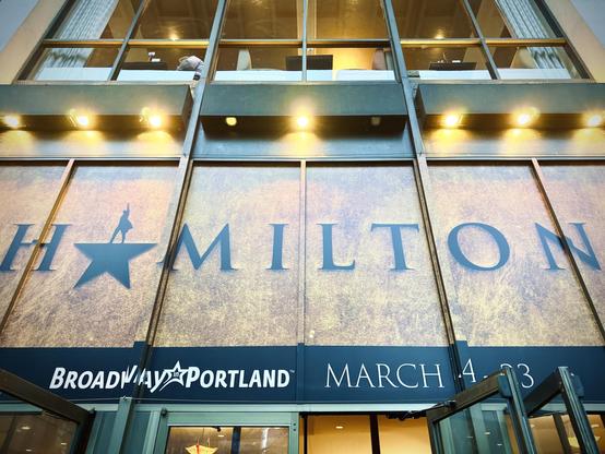 A large window banner in rust texture and black displays "HAMILTON," with white text over a black lower-thirds portion reading, "Broadway in Portland | MARCH 14 - 23."