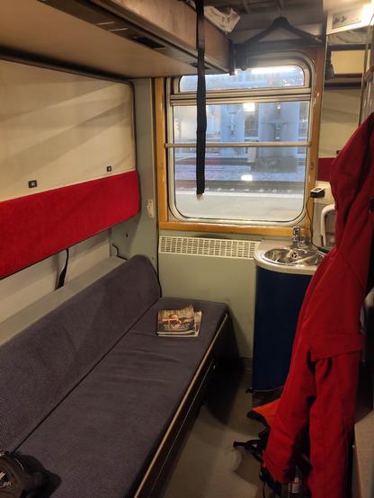 A three berth Swedish sleeper cabin, blue bench on the lower level, a mid-bunk packed away and above a higher level bed
 In the corner a small wash handbasin. A red coat hangs on a peg