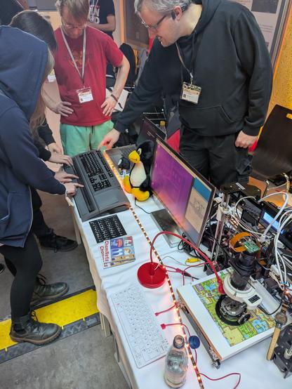 Der Stand des Chaostreff Chemnitz auf den Chemnitzer Linuxtagen. Zwei Standbetreuer und zwei Gäste schauen auf eine alte Braillezeile. Daneben steht ein selbstgebautes Mikroskop. Das Mikroskop ist ein alter 3D-Drucker an dem statt eines Druckkopfes eine Kamera mit einem Makroobjektiv montiert ist. In der Kamera wurde der Infrarotfiler ausgebaut. So kann das Mikroskop durch Silizium schauen. Auf einem Bildschirm ist das Innere eines Chips zu sehen.