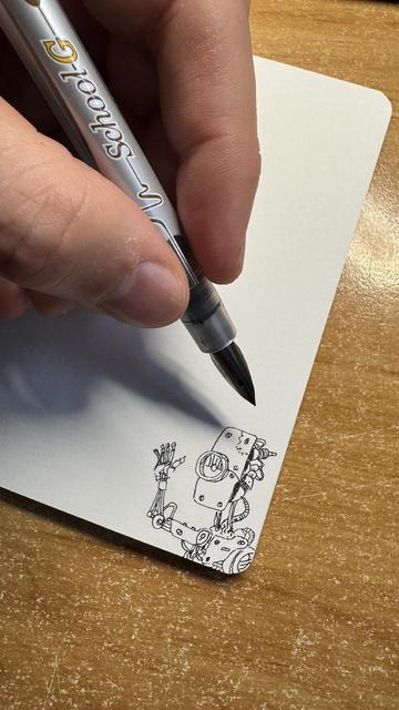 Photo of a hand with an ink pen drawing a robot on a smal sketchbook’s page corner. Robot is waving a hand and has a single eye piece.