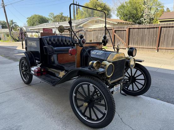 Three-quarter view of an early 20th century Model T Ford automobile originally designed to be an emergency response vehicle for police or fire. It is immaculately kept and fully functional, the brass trim and shiny black metal body with wooden frame pieces around the long black leather seat / couch in front if a very small truck bed that reads PATROL in gold lettering on the side.