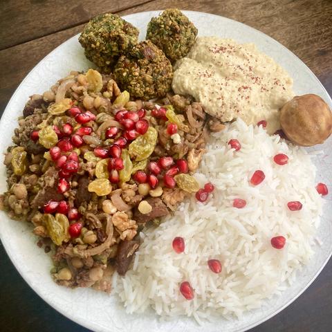 Overhead shot of a white plate with food. In a clockwise direction from the top, three falafels, a dollop of hummus dusted with sumach, a dried lime for presentation, a pile of basmati rice with a few pomegranate arils and finally a larger pile of the mung bean, split peas mix topped with yellow raisins and pink pomegranate arils