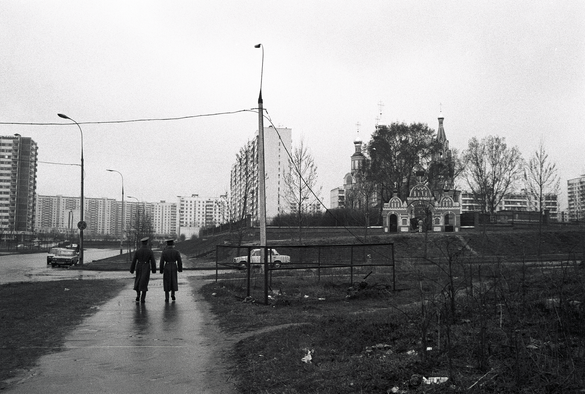 Two soldiers walking away from the camera on a sidewalk.  Highrises in the distance and a small church to the right of the frame, Russian Orthodox.
