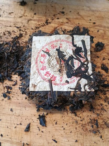 A clump of compressed tea with a square of paper stuck to it. The paper square has chinese characters on it and some individual tea leafs stuck on top of it.