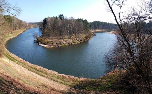 Schleife der Neiße im Panoramablick, vorne ein Trampelpfad, dahinter der blaue Fluss mit einigen Bäumen in der "Kurve", rechts und links noch Bewuchs. Am blauen Himmel keine Wolke.