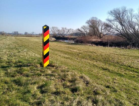 Bundesdeutscher Grenzstein, schwarz-rot-gelb gestrichen, vor einer Wiese  an der Neiße, im Hintergrund Fluss, Auen und Uferbewuchs