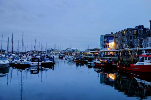 A tranquil evening scene at Sutton Harbour, Plymouth, England, with numerous yachts and fishing boats moored in the calm, dark blue water. The quayside is lined with buildings, some illuminated with warm lights reflecting on the water's surface. The sky is overcast, suggesting either dusk or dawn. One prominent red fishing vessel has the registration "PH819" clearly visible on its side.