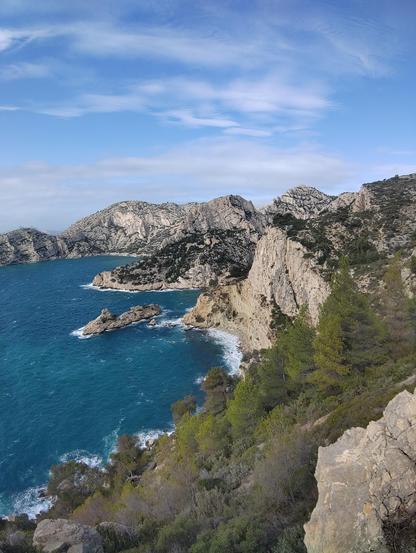 Vue depuis le chemin qui passe sous le socle de la Candelle.

Au première plan, on peut voir une pente pleine de végétation, avec arbustes et arbres.

On voit au deuxième plan la calanque des pierres tombées avec sa falaise verticale bien caractéristique. En face l'ile dite du "Torpilleur".

Au troisième plan, le cap Sugiton.

Au dernier plan le cap Morgiou, dont on ne voit pas le bout sur la photo.

La mer est bien bleu, presque turquoise. Et le ciel est aussi bleu avec son ton à lui. Quelques nuages blancs donne de la dimension à la photo.