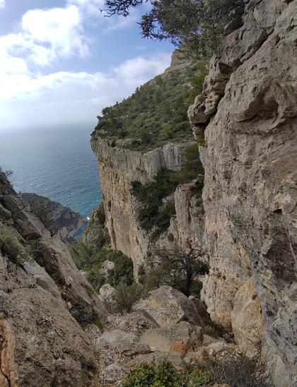 Après avoir passé le pas du la cheminée du diable, on se retourne et on voit bien le chemin impressionnant qui fraye son chemin dans une ligne de faiblesse évidente et large dans la falaise. Tout passe à pied, mais il faut bien les lever 😁. Un seul pas d'escalade en 3/4 facile mais un peu expo.

La photo montre en contre-plongée le chemin. Entouré de roche calcaire. 

On voit en contrebas une grande falaise bien verticale dont le sommet est chapeauté par un plateau en pente bien rempli de végétation.

Au loin, la mer s'étend à perte de vue et l'horizon est rempli de nuages blancs mais le ciel est bleu!