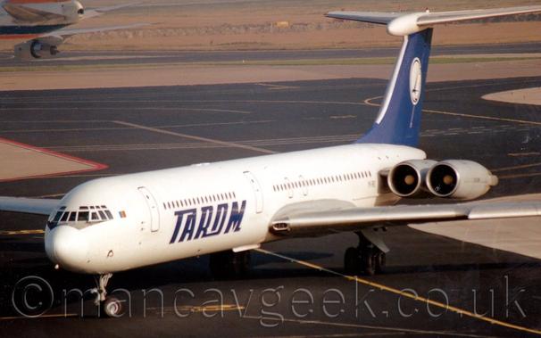 Side view of a white, 4 engined jet airliner with the engines mounted in pairs on the sides of the rear fuselage.
There are large white "Tarom" titles on the sides of the lower fuselage.
The tail is blue, with a large white circle in the middle containing a blue flying bird.
Black tarmac taxiways fill the foreground and most of the background, with wide strips of grey concrete marking the edges of the taxiways, often with grassed areas beyond the concrete.
The rear fuselage of a very large jet airliner with an orange tail can be seen in the top left corner, taxiing out of frame.