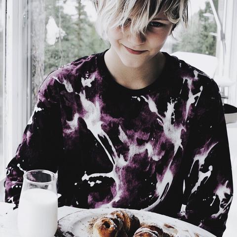 A child with tousled blond hair, wearing a vibrant purple and white abstract-patterned shirt, sits at a white table with a plate of cinnamon rolls and a glass of milk. A snowy forest is visible through large windows behind them.
