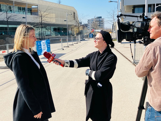 Das Bild zeigt drei Personen am Maria Trapp Platz vor der Baustelle. Eine Reporterin im schwarzen Mantel und mit Brille führt ein Interview mit einem knallroten Mikrofon mit ORF Aufdruck. Sie hört aufmerksam zu. Katarína von SeeStadtgrün, eine blonde Frau in einem dunklen Mantel erzählt etwas. Sie wird von einem Kameramann gefilmt. Der Himmel im Hintergrund ist blau, es sind auch noch einige kleine, kahle Bäume zu sehen.