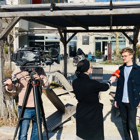 Das Bild zeigt drei Personen vor der Seestadt Lounge am Hannah-Arendt-Platz. Ein Mann filmt mit einer großen Kamera. Eine Frau im dunklen Mantel interviewt mit einem roten ORF Mikrofon einen Mann mit Brille und blauer Jacke. Hinter der Seestadt Lounge sieht man den Gastgarten des Restaurants See22. Dort sitzen einige Menschen mit Jacken, ein Fahrradfahrr fährt im Hintergrund vorbei.