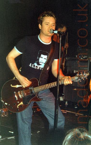 A man with short brown hair, wearing a dark blue t-shirt with a photo of Muhammed Ali on the chest and the word "Allstars" underneath, and a pair of blue jeans.
He is playing a brown guitar, and singing into a microphone on a stand in front of him.
In the background, a man in a red shirt is holding a guitar.
Black walls and ceiling fill the rest of the frame.