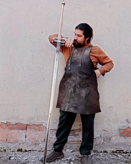 A swordsmith in a worn leather apron stands outside a building next to a schlachtschwert - a two-handed late medieval battle sword. The sword is nearly a foot longer than the smith is tall, and the smith is resting his raised arm on the rings between the handle and the blade.