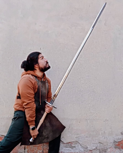 A swordsmith in a worn leather apron stands outside a building holding a schlachtschwert - a two-handed late medieval battle sword. He's holding the sword in a low guard, hands held low and sword point high. He looks up at the blade as if thinking "fuck, this is a really fucking long sword."