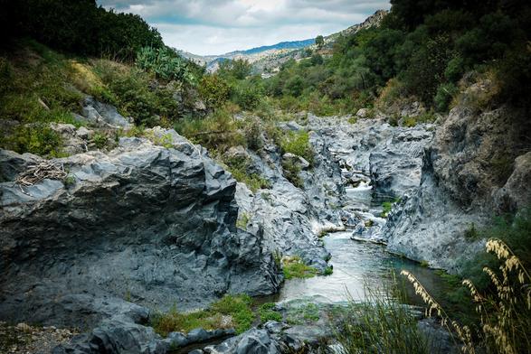 The image depicts a rocky riverbed with a small stream flowing through it. The river is bordered by large, dark gray rocks with a rough texture, creating a natural pathway for the water. The water appears clear and is moving gently, with small ripples visible in some areas. The surrounding landscape is lush with green vegetation, including shrubs and grasses, which grow on the rocky terrain. In the background, rolling hills are visible under a cloudy sky, adding depth to the scene. The overall color palette is dominated by shades of gray from the rocks, green from the vegetation, and blue from the water, creating a serene and natural setting.

Provided by @altbot, generated privately and locally using Ovis2-8B