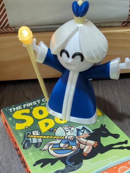 3D printed "Moon Queen" comic character in a blue dress holding a yellow staff that's lit up. Sitting on top of the book "The First Cat In Space and the Soup of Doom"