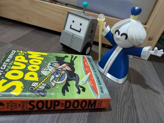 3D printed "Moon Queen" comic character in a blue dress holding a yellow staff that's lit up. Sitting next to Loz 4000, a gray 2-wheeled boxy robot, also printed. Foreground is the book "The First Cat In Space and the Soup of Doom"