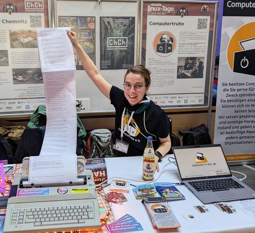 Bild zeigt Karola am Stand des ChaosTreffs und der Chemnitzer Computertruhe. Dabei hält Karola mit dem linken Arm das bedruckte Endlospapier hoch und im Vordergrund sieht man den Merch und Monitor der Computruhe .Im Hintergrund sind die Poster und Aufsteller.