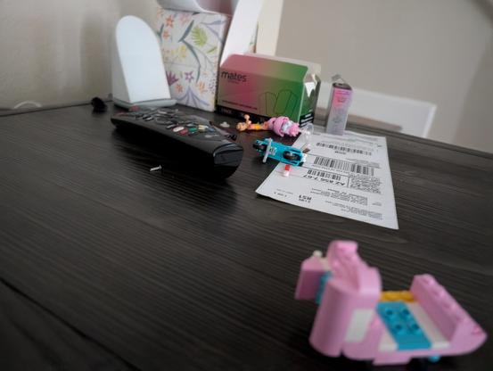 A partially assembled Lego Friends cotton candy stand sits on a dark wooden end table. The structure is built on two 2x6 plates, which are connected by a few tiles and jumper plates for added stability. The stand features a playful color scheme with pink, blue, and yellow bricks. Two 1x4 plates with pins extend from the bottom, intended for wheel attachment in a later stage of the build. The design includes several pink 1x2 and 2x2 bricks forming the main structure, along with curved bricks and sloped pieces that create a rounded, whimsical appearance. .