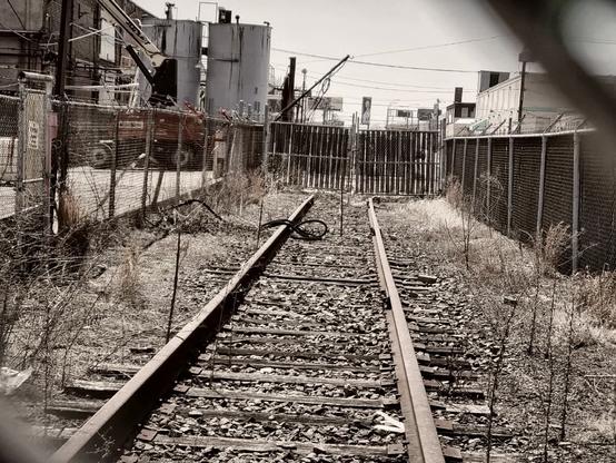 Black and white photo of abandonded train tracks leading to a chainlink fence. The photo was taken through the fence. Portions of the chainlink can be seen at the edges of the photo. Weeds and grass grow up through the rails and ties. Multiple industrial structurea can be seen in the distance on the right. The overall feel is cold and ominous.