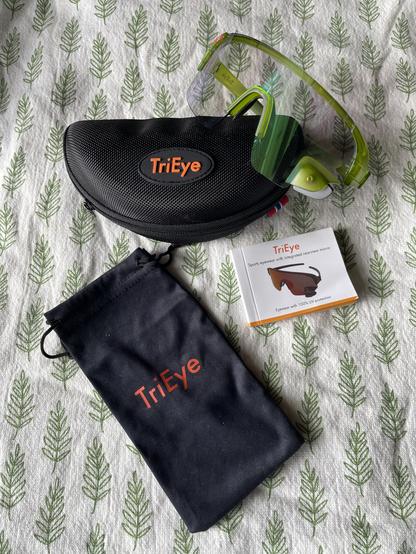 A green pair of Tri-Eye photochromatic cycling glasses with mirror is perched alongside its soft sleeve and hard case—both black with the word TriEye in orange—and small TriEye product manual.