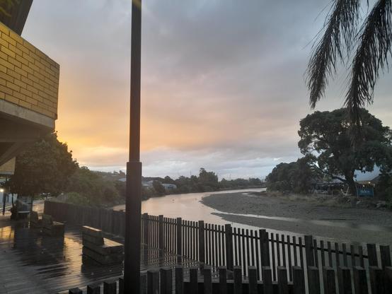 The sky is beginning to turn yellow and orange in a sunset. The boardwalk is visible with some bench chairs, and the wooden surface is shiny because it's wet from rain. It looks out to the Maitai River which is not very full currently.
