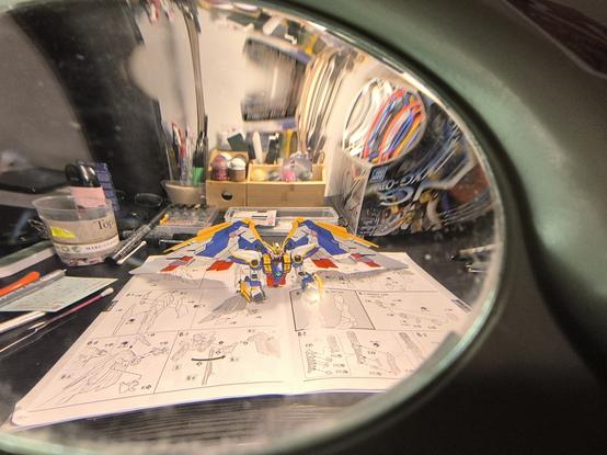 Partie supérieure du gunpla terminée et présentée au travers de la lampe loupe avec ses deux grandes ailes (blanc, rouge, bleu et jaune). Il repose sur le livret de montage à la page des ailes et des jambes.