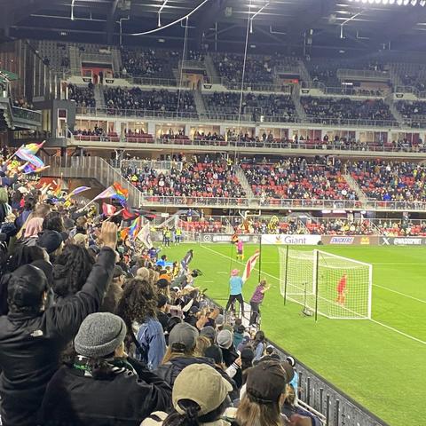 The mostly-full stands of Audi Field with the goalie in red and the fans behind waving DC, progress pride, rainbow, pansexual, and transgender flags among others.