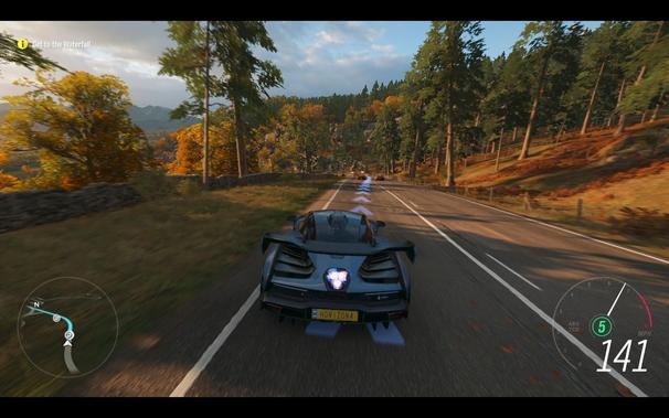 The player is in a car, driving on a curvy road through a forest. The road has yellow lane markings and the trees have golden leaves indicative of autumn season. The speedometer in the bottom left corner shows 141 mph. The dashboard displays a heart symbol with an exclamation mark inside it, indicating some sort of warning or alert for the player to pay attention to.